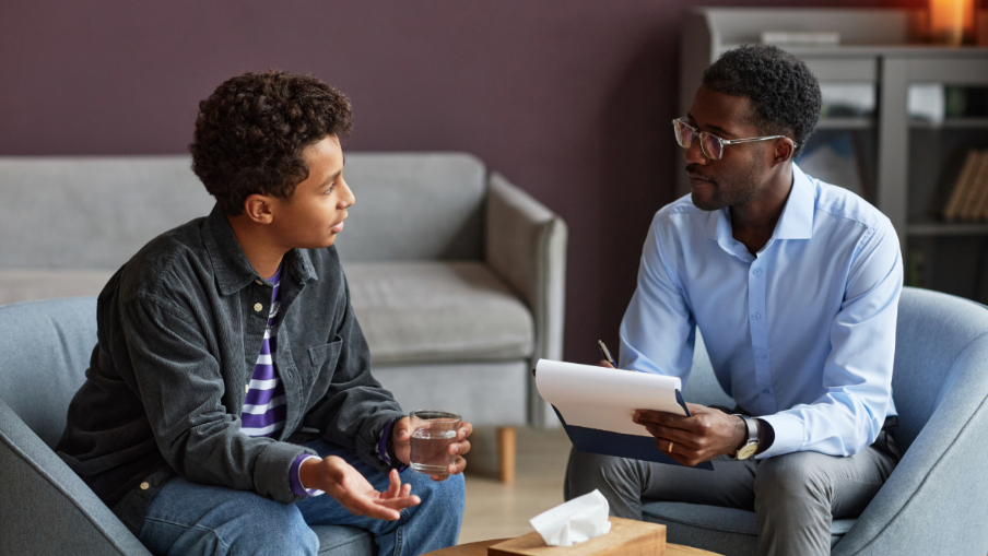 two men sitting and discussing, one is holding a glass of water and the other has glasses and is writing on a notepad