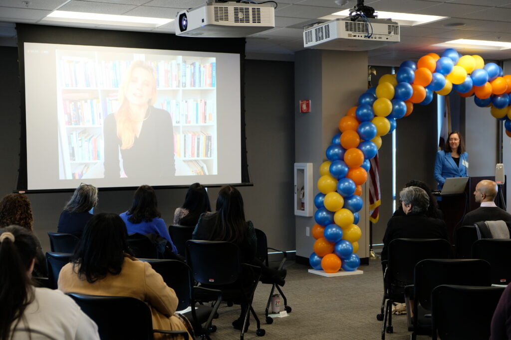 Department of Health Care Access and Information Director Elizabeth Landsberg Stands at podium listening to California First Partner Jennifer Siebel Newsom on the screen to the left. 